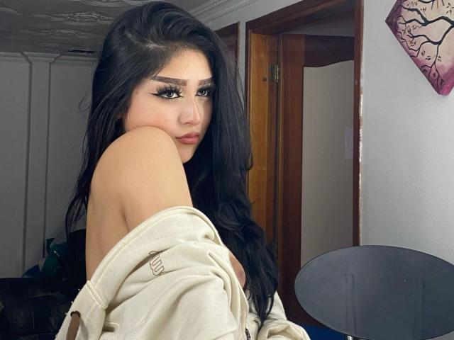 LilaRoux with long black hair, off-shoulder beige shirt, and thick black eyeliner, looking over her shoulder, showing her bare shoulder and upper chest, with a slightly pouty face.