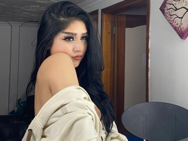 LilaRoux with long black hair, heavy eyeliner, and off-shoulder beige top, showing her bare shoulder and looking over her shoulder, with a slight pout. Her tits are partially visible.