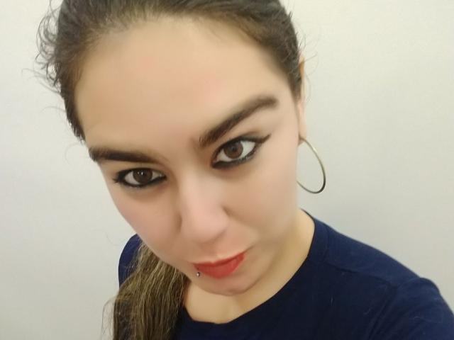 TaylorNova heeft donkere wenkbrauwen, zwarte eyeliner, rode lippen en een kleine piercing in haar onderlip. Ze draagt een blauw shirt en grote oorringen, en haar haar zit in een paardenstaart..
