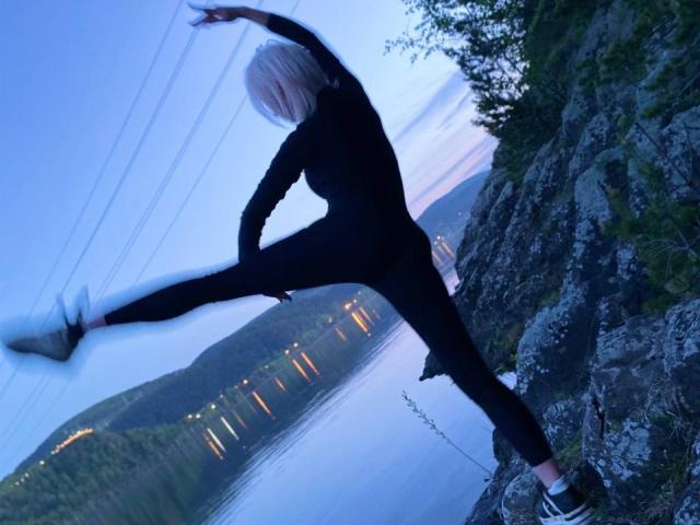 BlondIce está realizando una patada alta en un acantilado rocoso, vistiendo un traje de cuerpo entero negro y zapatillas deportivas, con una iluminación crepuscular azulada y una vista reflectante del agua detrás..