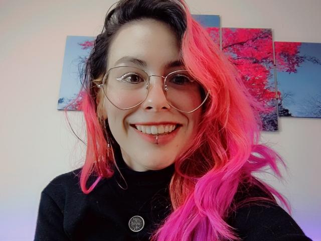 NatashaLau, com cabelo rosa e preto vibrantes, usando óculos redondos e uma blusa preta, sorrindo com um piercing no septo e um piercing no lábio, exibindo seus dentes atraentes..