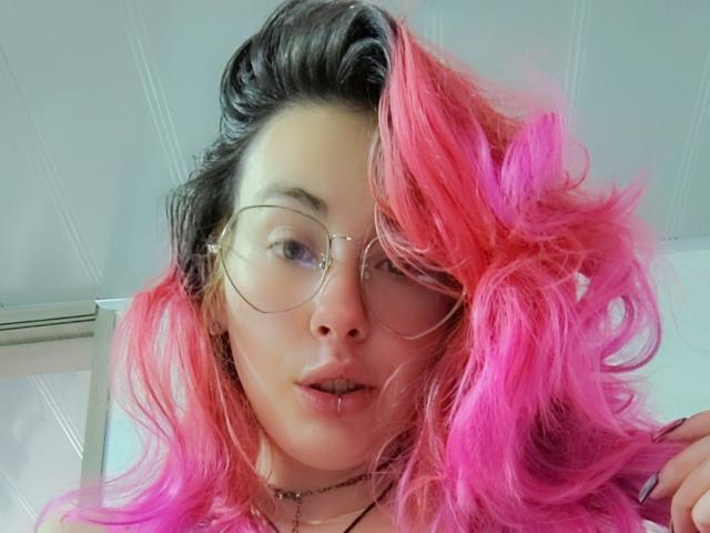 NatashaLau with pink and black curly hair, wearing glasses, has a septum piercing and a necklace, looking at the camera with a slight pout, holding her hair.