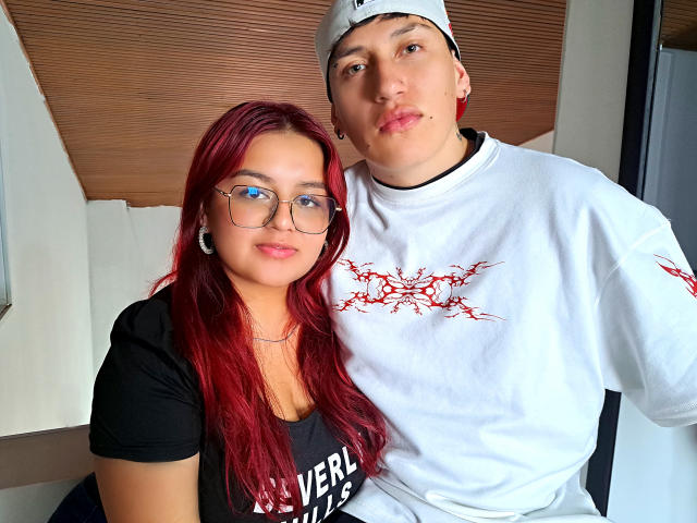 JaneAndLeo mit roten Haaren und Brille, in einem schwarzen Oberteil und mit kleiner Brust, steht neben Leo, der ein weißes Hemd, rote Tattoos und einen schlaffen Gesichtsausdruck hat und eine Mütze trägt..