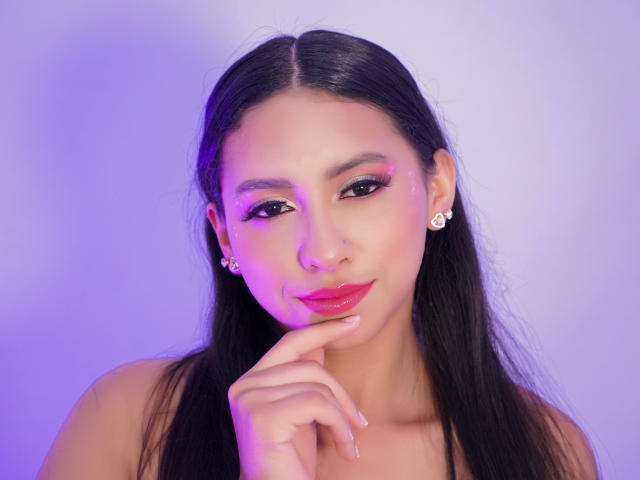 AshleyRosel with long black hair, pink eye shadow, red lipstick, nose ring, earrings, and finger touching her lips, smiling, purple-blue lighting. Boobs not visible.