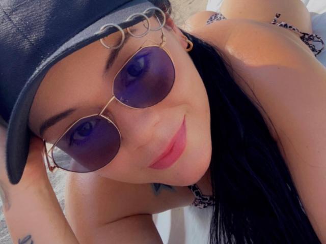 ChristyCuteX with purple-tinted aviator sunglasses, black cap, and pink lips, lying on a white towel, showing a tattoo on her arm, with black hair and a smile.