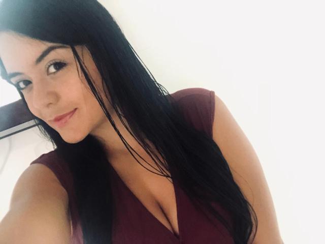 ChristyCuteX with long black hair, brown eyes, and a slight smile, wearing a deep burgundy V-neck, showing noticeable cleavage and medium-sized tits.