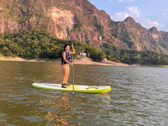 ChristyCuteX stands on a green paddleboard, wearing a blue life jacket and black shorts, with a paddle in hand, on a calm, reflective lake with rocky mountains.