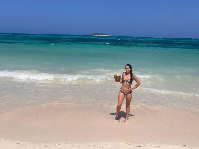 ChristyCuteX stands on a sandy beach holding a coconut, wearing a leopard print bikini, with blue-green ocean waves and a small island in the background.
