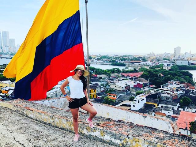 ChristyCuteX with a yellow, blue, and red flag, white shirt, black shorts, pink sneakers, and a sunhat, standing on a rooftop, city skyline in the background.