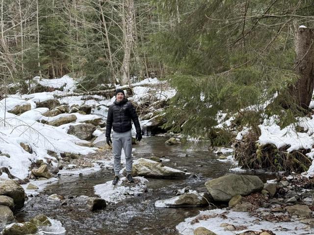 HarrisonSun stands in a snowy forest creek, wearing a black jacket, gray pants, and boots, with a bearded face and a backpack, surrounded by rocks and green pine trees.