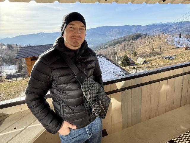 HarrisonSun with a black beanie, puffy black jacket, and checkered bag, standing on a wooden balcony, hands in jeans, mountains and clear sky in the background.
