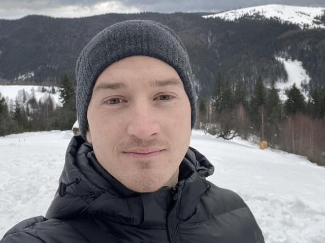 HarrisonSun, con un berretto grigio e un giubbotto imbottito nero, ha il viso rasato, la pelle chiara e gli occhi marroni, ed è in piedi in un paesaggio montano innevato..
