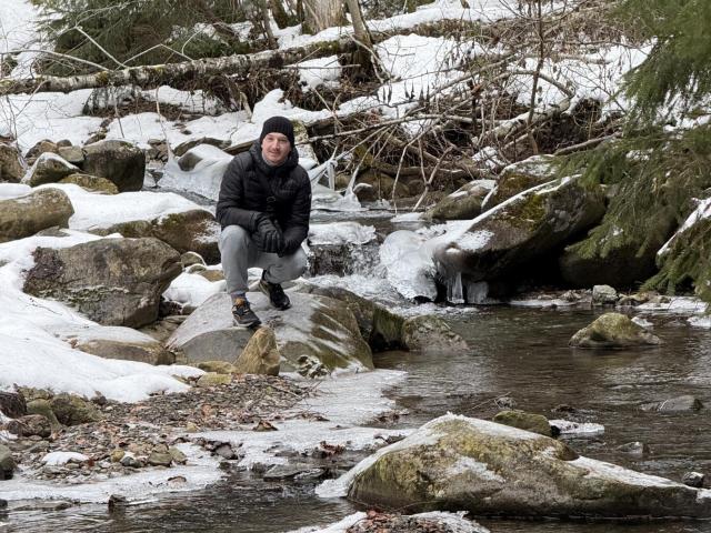 HarrisonSun è seduto su una roccia coperta di neve, indossa un berretto nero, una giacca nera, pantaloni grigi e guanti neri, con un ruscello e rocce ricoperte di muschio sullo sfondo..