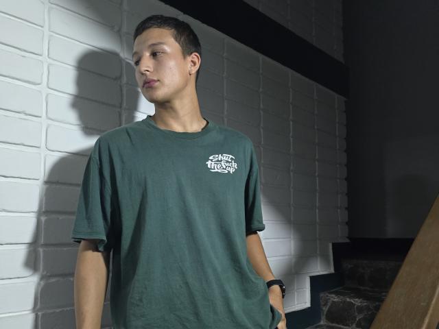 DavidLatinX stands in a dark room wearing a green t-shirt, hands in pockets, shadow on white brick wall, black wristband, short dark hair, looking to the side.