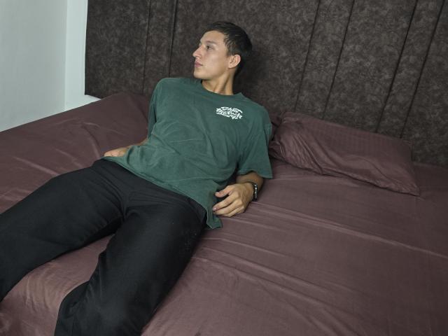 DavidLatinX is lying on a bed, wearing a green shirt and black pants, with a relaxed, slightly upward gaze, hands resting on his stomach, dark textured headboard behind.