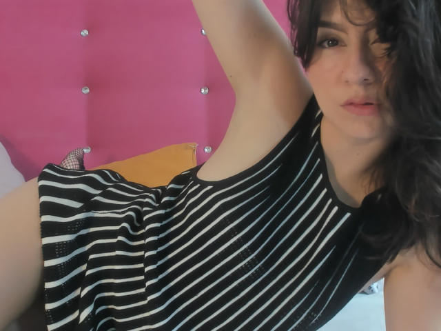 Kari Carter, met golvend zwart haar, draagt een zwart-wit gestreepte jurk, met een arm omhoog, waarbij haar oksel zichtbaar is, en kijkt naar de camera met licht open lippen. De achtergrond is een roze muur met textuur..