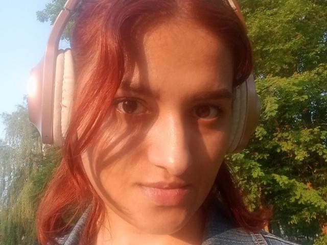 KathleenJenny with red hair, wearing large beige headphones, has a serious expression, and a light skin tone, standing outdoors with green trees in the background.