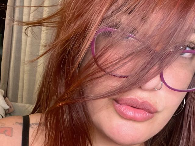 ReinaMomoo's rode haar bedekt haar paarse bril, en je kunt een neusring en roze lippen zien. Ze heeft tatoeages op haar schouder en draagt een zwarte band. Haar ogen zijn gedeeltelijk zichtbaar..