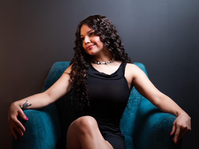 NatallyFlame with curly black hair, wearing a black dress and a necklace, sitting in a teal chair, smiling, arms resting on arms, tattoo on right wrist, pink nails.