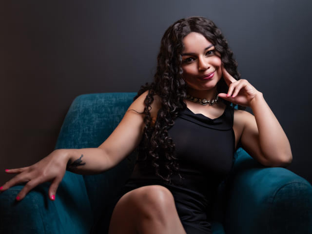 NatallyFlame, with curly black hair, black dress, and necklace, sits in a teal chair, touching her chin with a finger, showing a tattoo on her left hand.