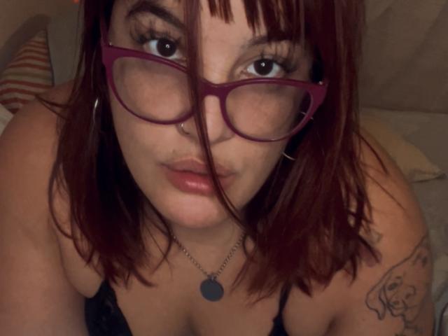 ReinaMomoo con i capelli rossi, gli occhiali viola, il piercing al naso e gli orecchini a cerchio, che guarda la fotocamera, mostrando il décolleté con una maglia nera in pizzo, un tatuaggio sulla spalla destra e una collana con un ciondolo rotondo..