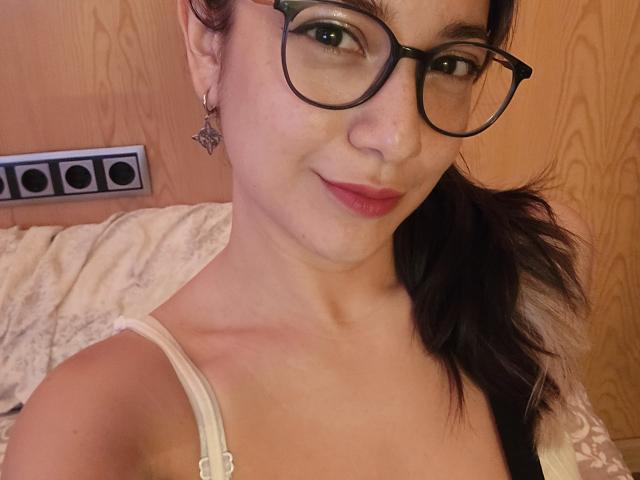ErinFoxy with glasses, pink lipstick, and a slight smile, showing her tits in a white strap, dark ponytail, and a star earring, standing in a wooden room.
