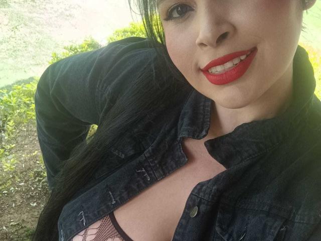 AgataP with red lipstick, black denim jacket, and visible cleavage in a fishnet top, smiling at the camera, outside in bright greenery.