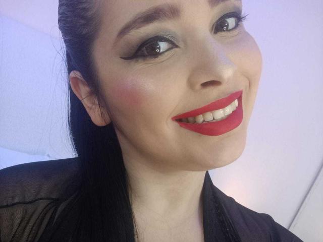 AgataP has red lipstick, black eyeliner, and a smile showing white teeth. She's wearing a black sheer top, with black hair pulled back, looking at the camera.