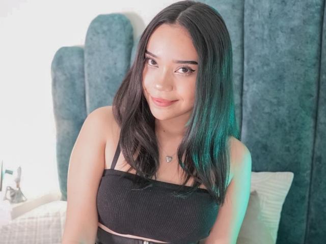 MarylinLewis has dark, straight hair and a cute smile. She's wearing a black, ribbed, strapless top that highlights her small tits. She's sitting in front of a blue headboard.