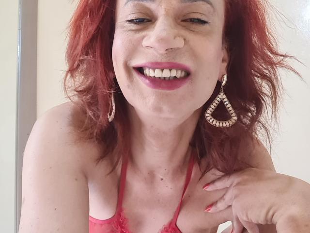 BrunelaSwan with red hair, smiling, wearing pink lace top, teardrop earrings, and pink lipstick, has nose ring and red nail polish, showing her tits slightly.