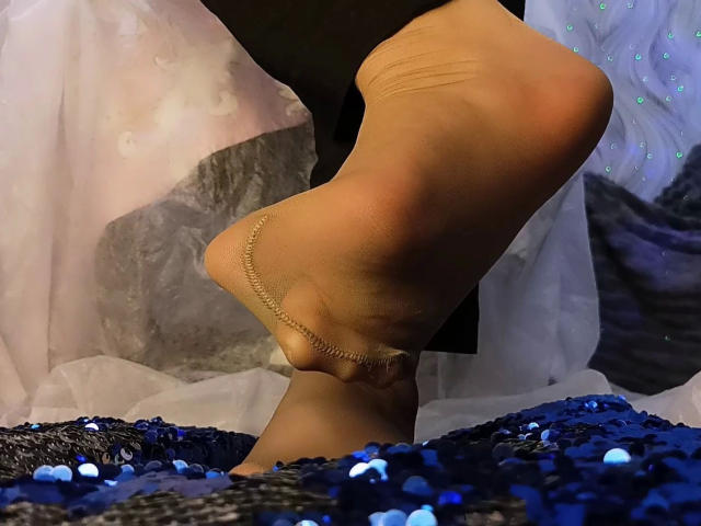 Rachel-Sweety-hot's foot in a sheer, tan-colored sock with a gold ankle chain, standing on a blue, sequined surface. Her heel is slightly raised, showing her sole.