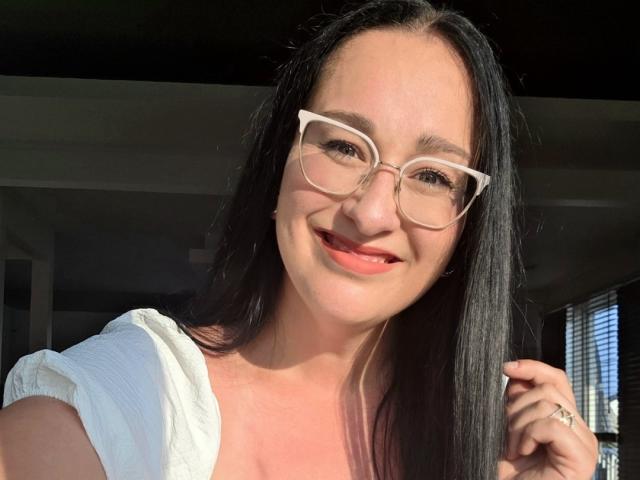 RoriQ has black hair, white glasses, and is smiling with red lipstick. She's wearing a white top, sunlight on her face, and has a ring on her right hand.