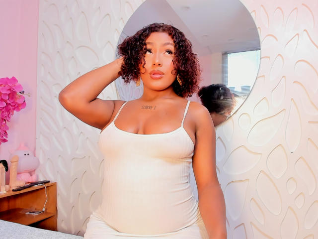 AmberQueeny with curly brown hair, wearing a white tank top, showing her tits, has a tattoo on her chest, and sits in a pink room with a floral mirror.