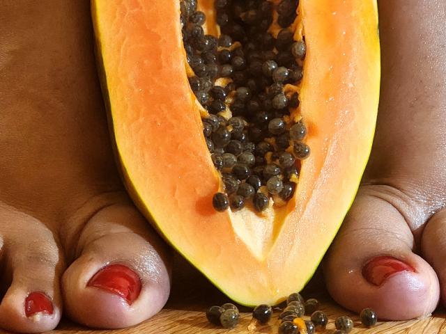 ViickyLee's feet with red toenail polish, holding a halved pumpkin with black seeds in front of her toes. The pumpkin's bright orange contrasts with her dark skin.