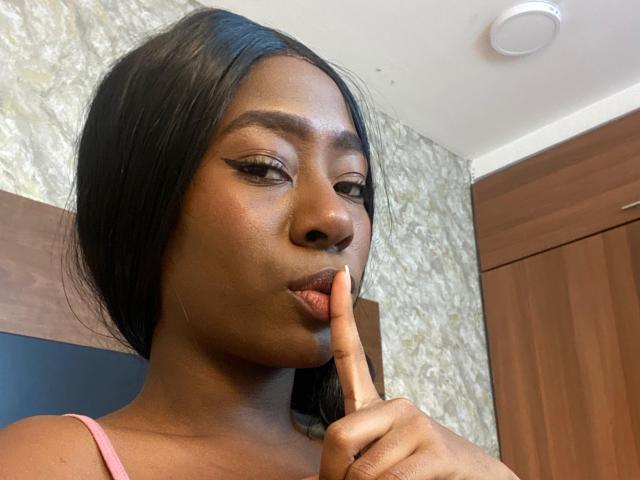 VioletGraice with dark skin, long black hair, and pink lips, holding a finger to her mouth, wearing a pink top, with winged eyeliner, looking slightly to the side.