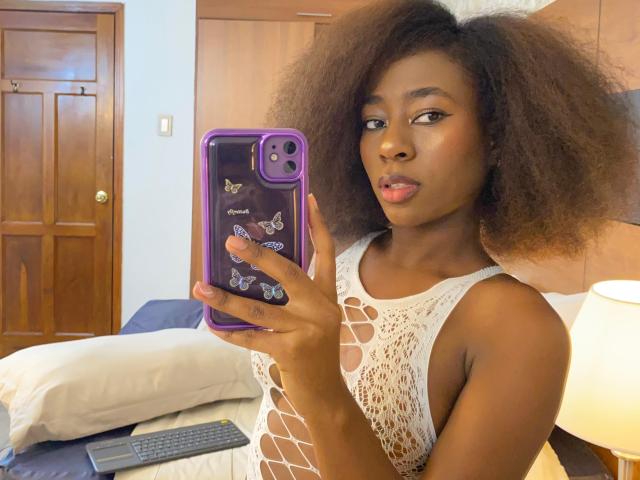 VioletGraice with dark brown skin, curly hair, and white lace top, holding a purple phone with butterfly decals, looking at the camera with pouty lips. Bed and lamp in background.