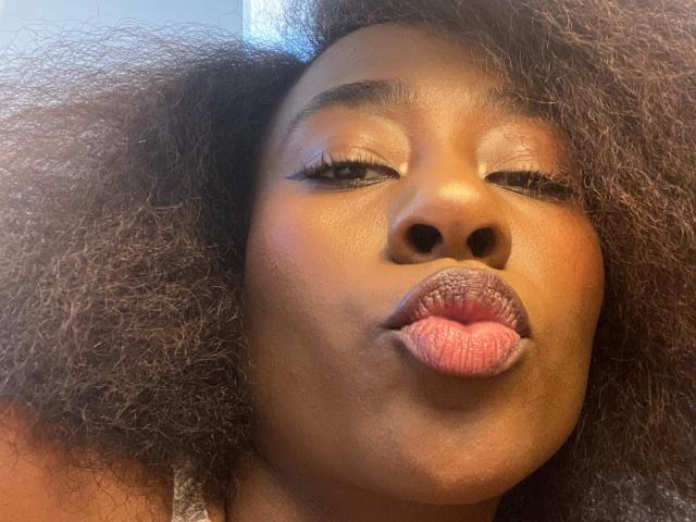 VioletGraice's face with dark brown skin, curly black hair, closed eyes, and puffy pink lips in a kissy face expression, showing a subtle sheen on her skin.