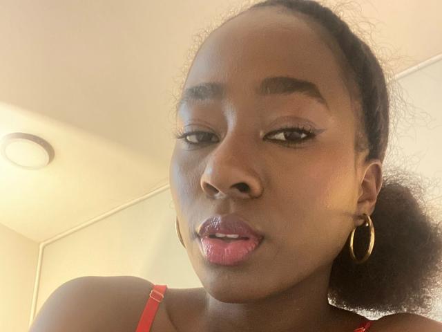 VioletGraice with dark skin, pink glossy lips, gold hoop earrings, red bra straps, and black curly hair, looking slightly to the side, with a subtle smile.
