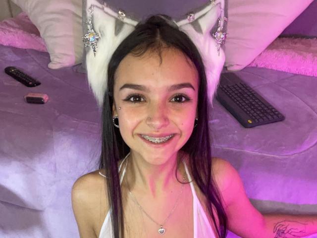 MarietteJones with cat ears, smiling, showing metallic teeth, white tank top, black hair, purple light, remote, phone, and keyboard on bed. Boobs are not visible.
