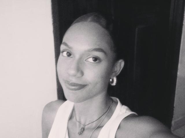 AylaCallowey has a smooth, dark-skinned face with a slight smile, wearing a white tank top, small earrings, and a necklace, in a black-and-white selfie.
