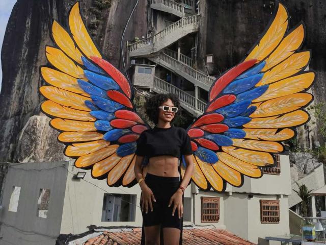 AylaCallowey with large, colorful angel wings (yellow, blue, red) wearing a black crop top and black shorts, standing with hands on hips, wearing white sunglasses.