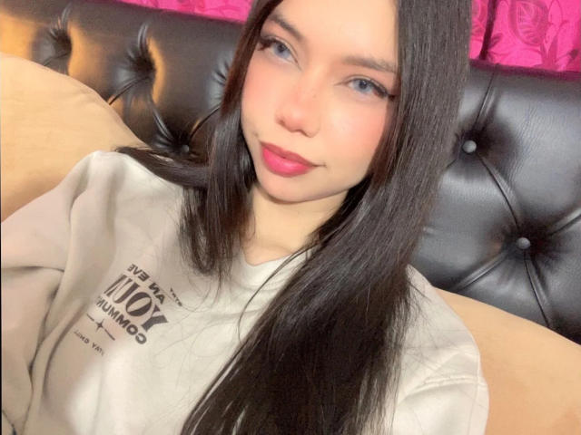 MiAntonella with long black hair, blue eyes, pink lipstick, and white sweatshirt, sitting on black tufted leather couch, smiling slightly, with beige pillow behind her.
