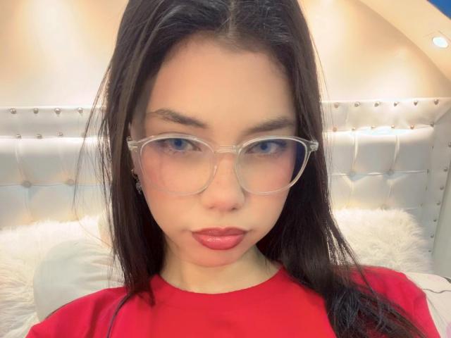 MiAntonella with long black hair, clear glasses, red lips, and red shirt, looking at camera with slight pout, on white bed with tufted silver headboard.