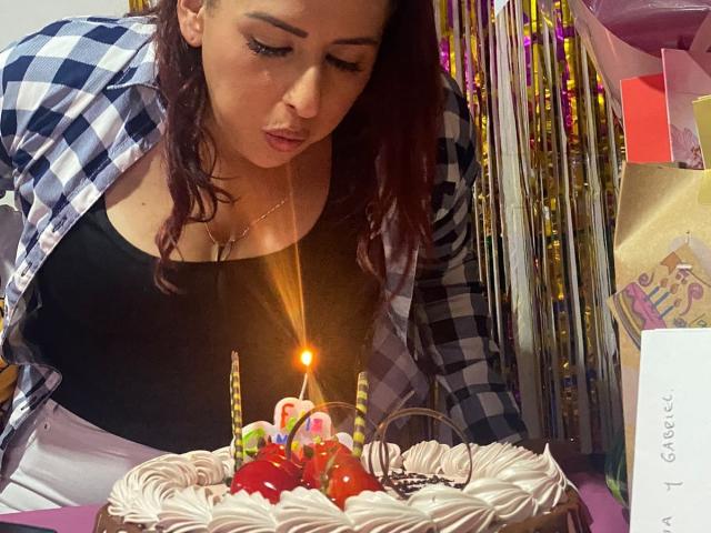 DylaraCollins with medium tits, wearing a black top and checkered shirt, blowing out candles on a cake with strawberries and whipped cream, in a celebratory setting.