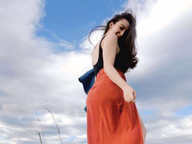 MabyLegrand with long dark hair, black tank top, and red skirt, holding a blue pillow, looking back with a slight smile, under a bright blue sky with white clouds.