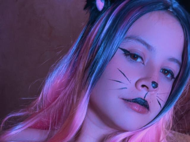 Melani Becker mit Katzenohren, schwarzer Nase, schwarzem Eyeliner, schwarzem Lippenstift, rosa und blau gefärbtem Haar, künstlichen Schnurrhaaren und einem leichten Lächeln, die in die Kamera schaut..