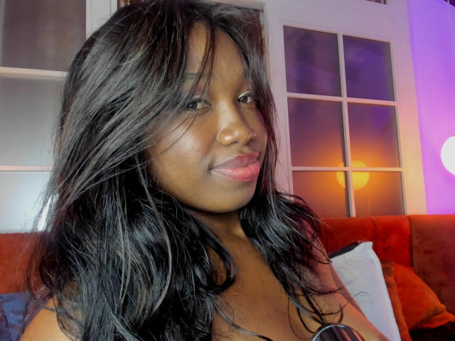 KarlieMoss with dark, wavy hair, medium brown skin, pink lips, and a subtle smile, showing her bare shoulders and a hint of her tits in a cozy setting.