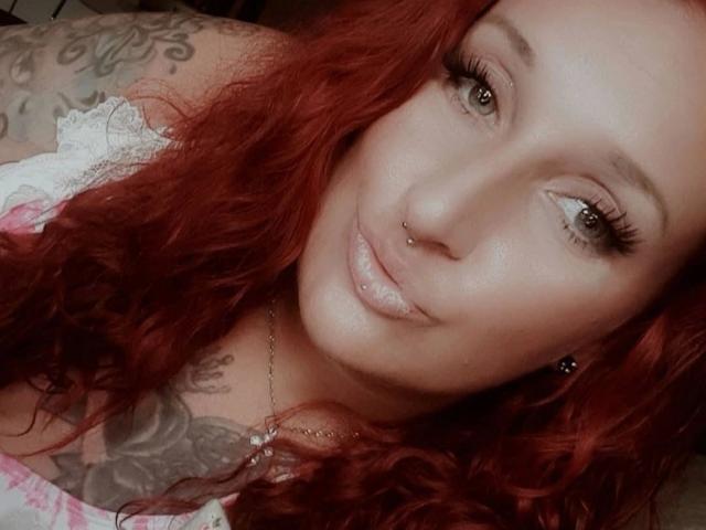 Licaink, con i capelli rossi ricci, un piercing al naso, tatuaggi sulle spalle, indossa una camicetta bianca in pizzo, ha labbra rosa lucide e lunghe ciglia, guarda la fotocamera con un leggero sorriso..