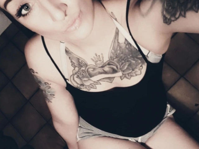 Licaink, con tatuaggi, indossa una canotta nera e pantaloncini grigi, mostrando un tatuaggio sul petto con ali e rose, e un piercing al naso, seduta su un pavimento piastrellato..