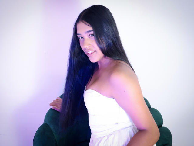 IvonneLaurent with long black hair, smiling, wearing a strapless white dress, sitting on a green chair, showing her tits, with a purple-blue light effect on her skin.